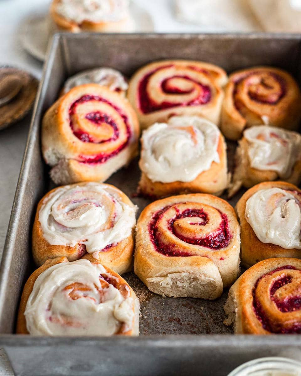 Jam Donut Cinnamon Rolls in baking tray. Some are iced and some are not. One roll is removed showing the feathery and soft edge of one of the rolls.