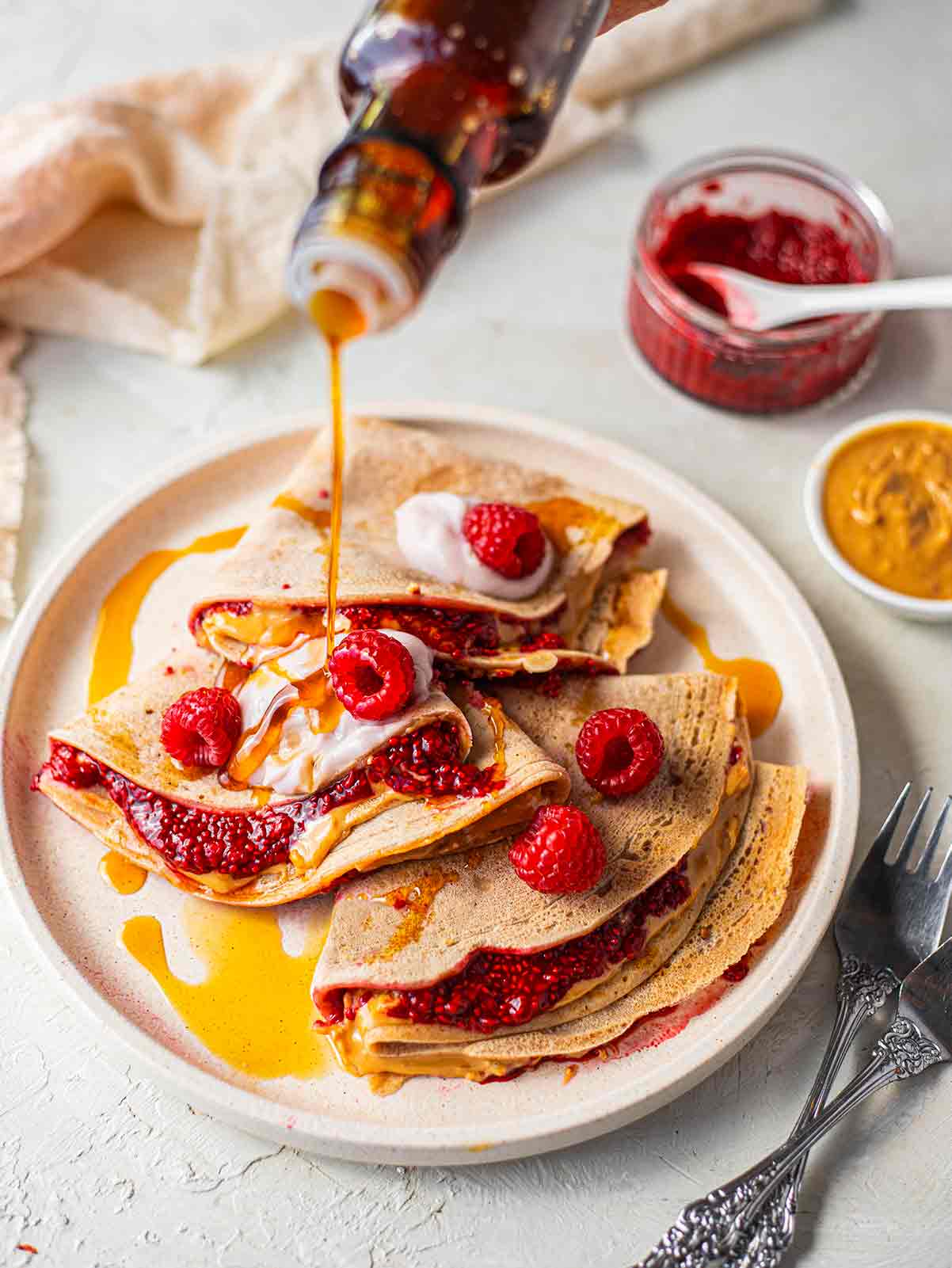 Stack of crepes filled with raspberries and peanut butter, topped with coconut yoghurt and drizzle of maple syrup.