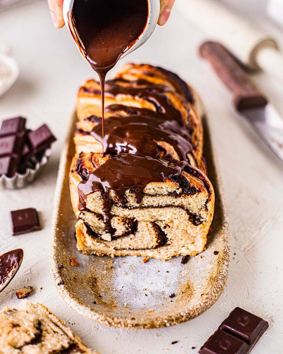 Vegan chocolate babka on serving platter. Babka has shiny chocolate ganache poured on top.