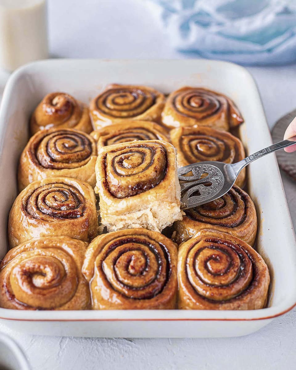 A dozen of vegan sticky cinnamon buns in baking tray with one centre bun lifted out with a cake server exposing feathery and soft edges.