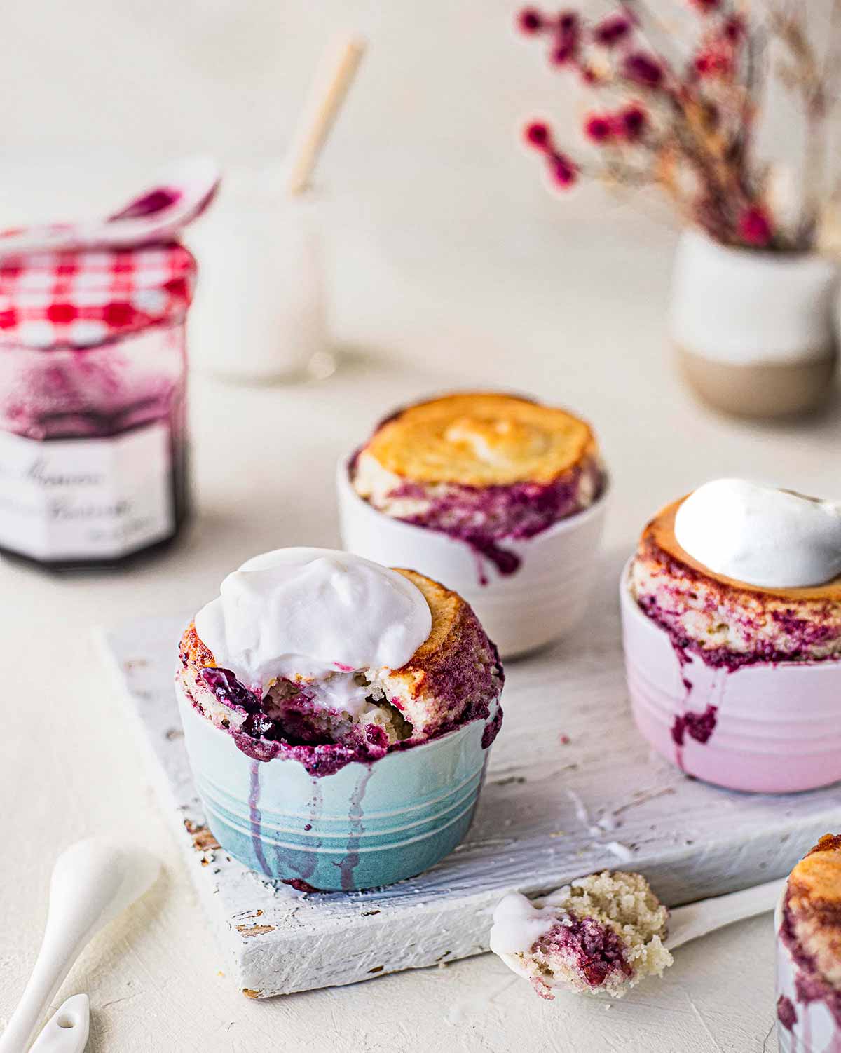 Self saucing puddings in little ramekins on white chopping board. Each pudding is bursting with jam on the sides and some are topped with yoghurt.