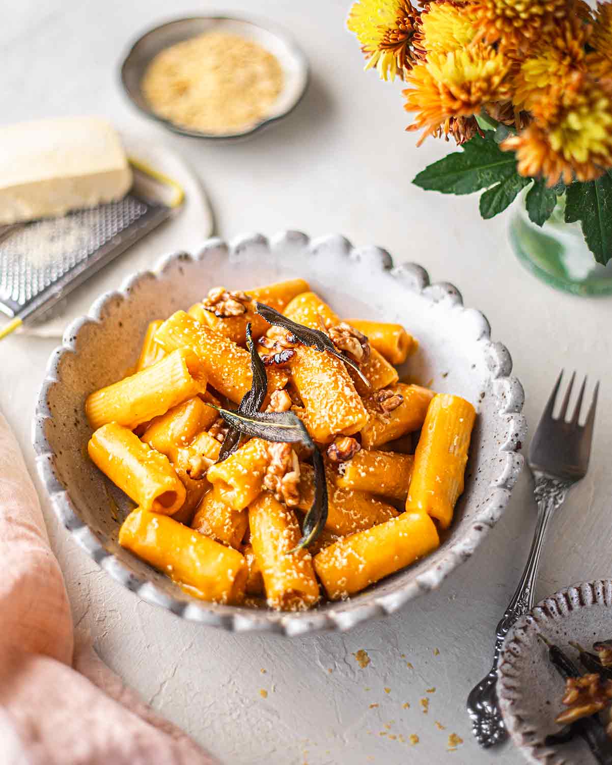 Vegan roasted pumpkin alfredo topped with sage leaves, walnuts and shaved vegan cheese in a bowl next to yellow orange flowers.