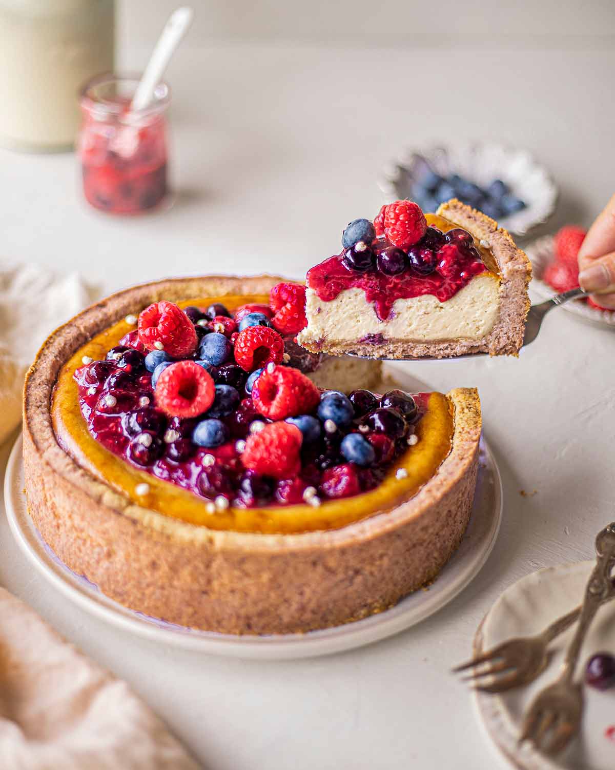 Vegan baked cheesecake topped with berries. A slice has been cut out and is lifted up by cake server to reveal creamy texture of cheesecake.