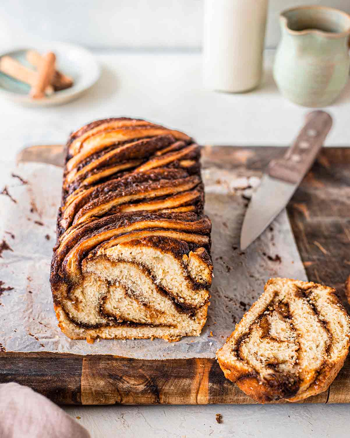 Vegan Cinnamon Babka with slice cut off showing interior. Babka is on a rustic piece of baking paper and a chopping board with a knife.