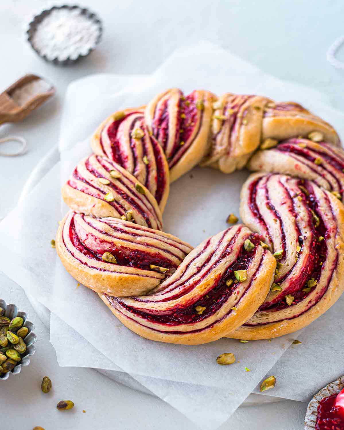 Close up of raspberry wreath with chopped pistachios on baking paper and marble serving board.