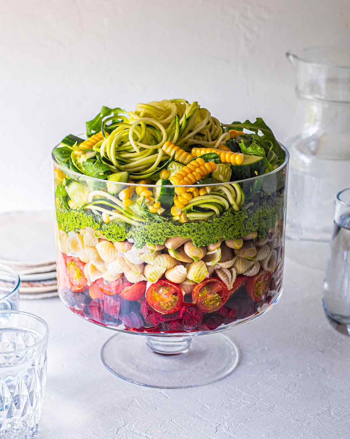 Vegan Layered Pasta Salad with Pesto in trifle glass with serving jugs and water surrounding.
