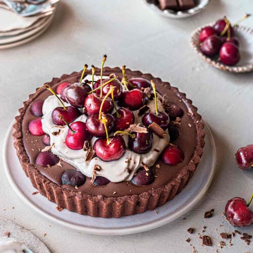 Vegan Blackforest tart with a chocolate base, ganache filling, coconut cream topping and lots of cherries on top. Tart is on plate surrounded by more fresh cherries and serving plates