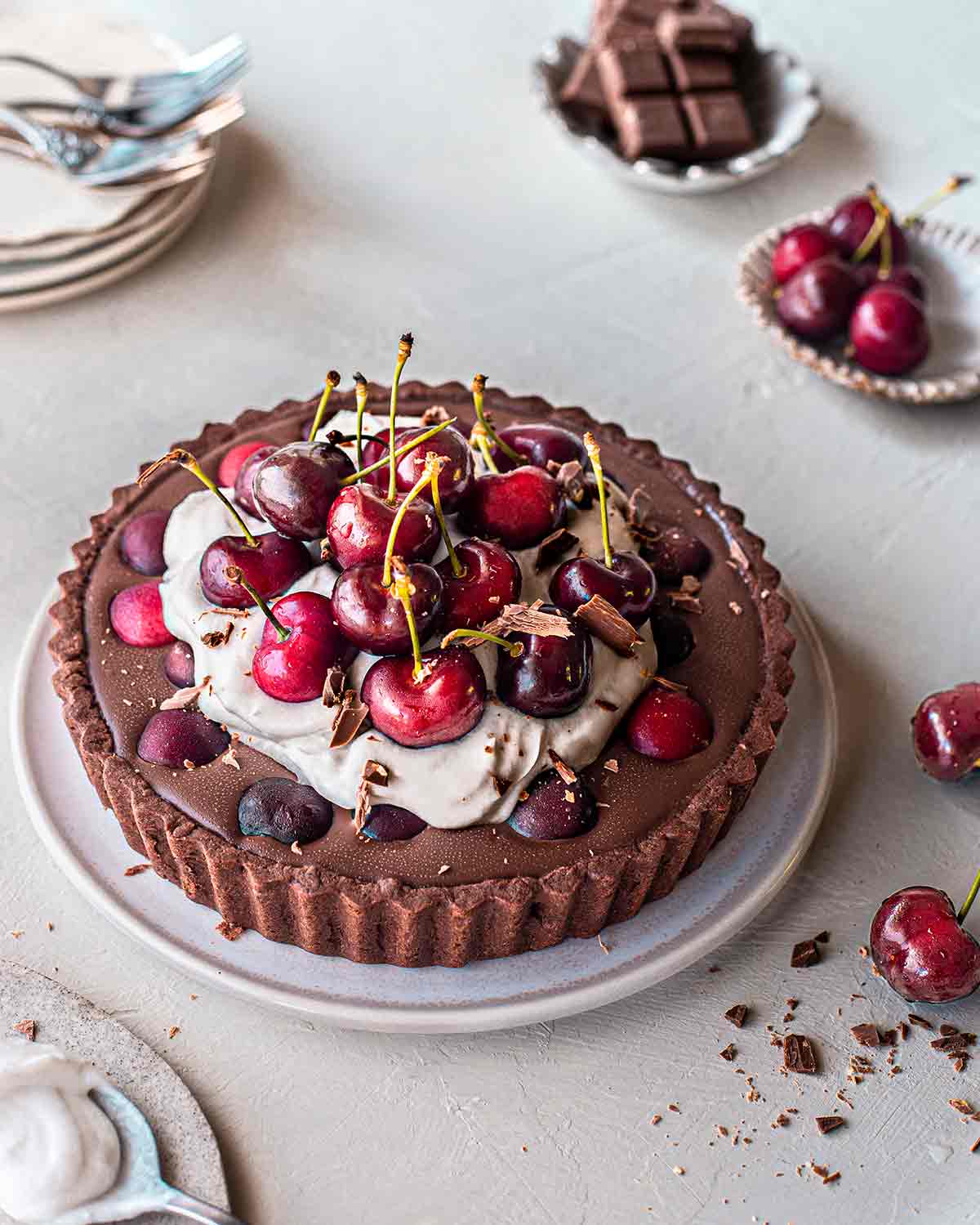 Vegan Blackforest tart with a chocolate base, ganache filling, coconut cream topping and lots of cherries on top. Tart is on plate surrounded by more fresh cherries and serving plates