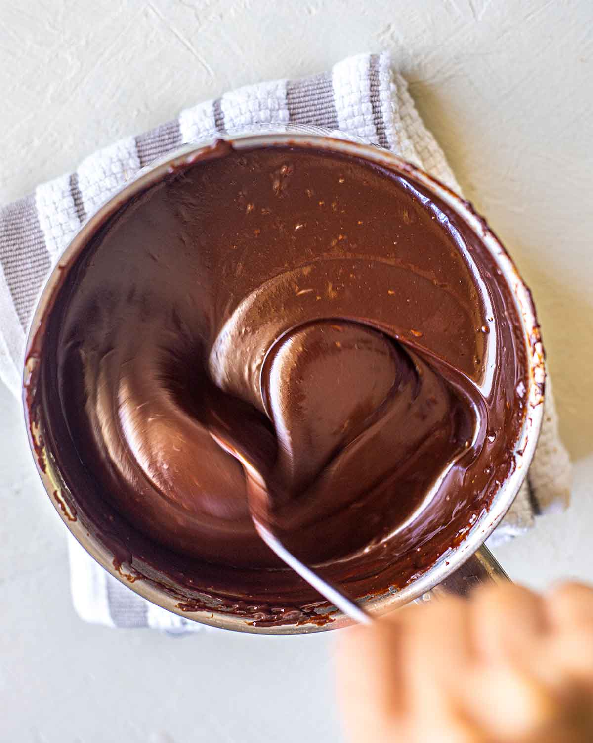 Close up of small saucepan with melted chocolate ganache. A hand is stirring a spoon in the ganache showing its thick consistency.