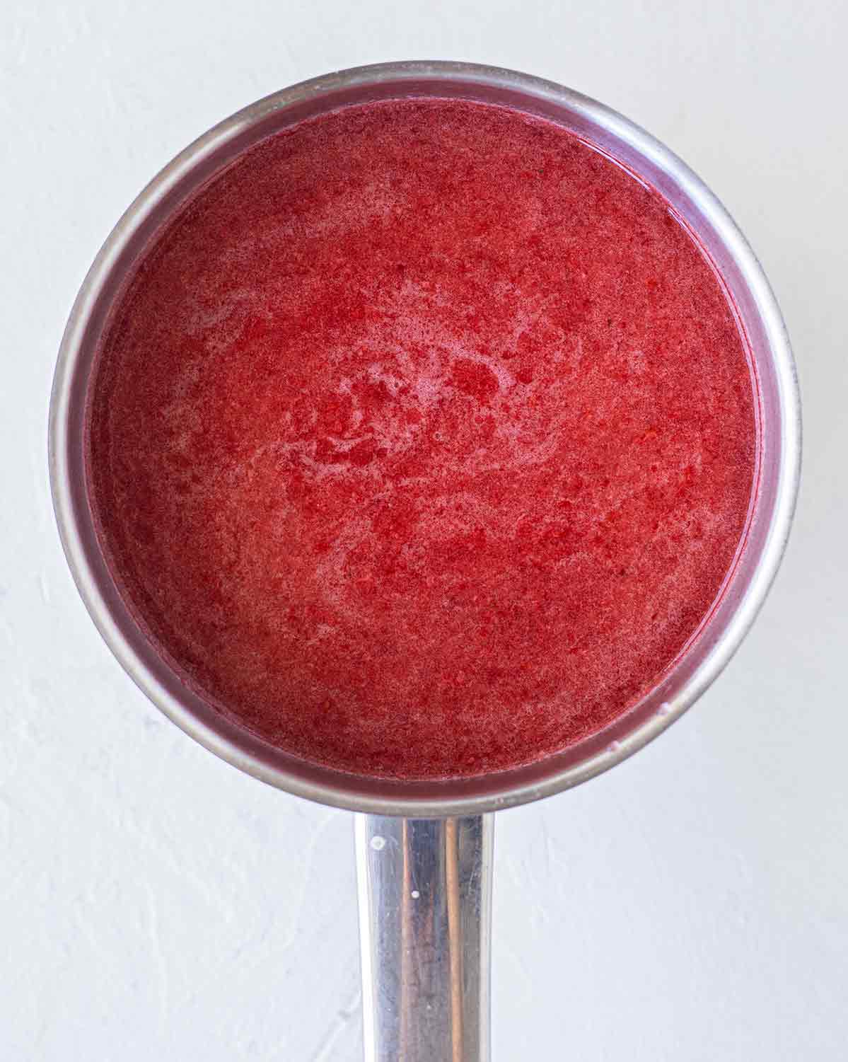 Close up of vegan jelly mixture in a saucepan. The mixture is an opaque red liquid with little bubbles throught.