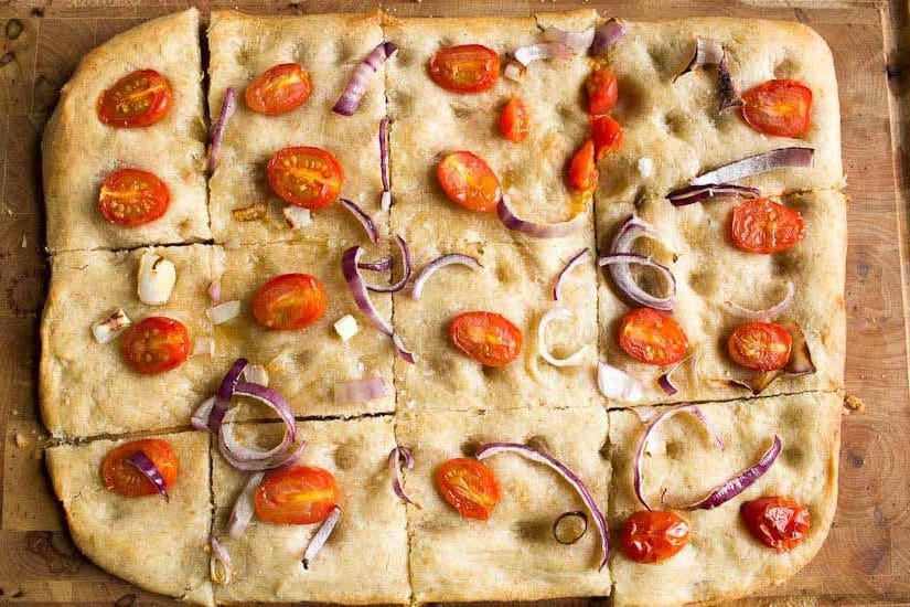 Sourdough focaccia with chopped cherry tomatoes and red onion slices cut into segments on chopping board.