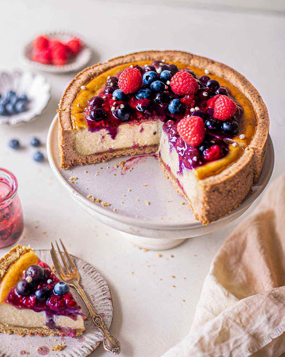 Healthy baked cheesecake (vegan) topped with berries on cake stand with cake slice in foreground.