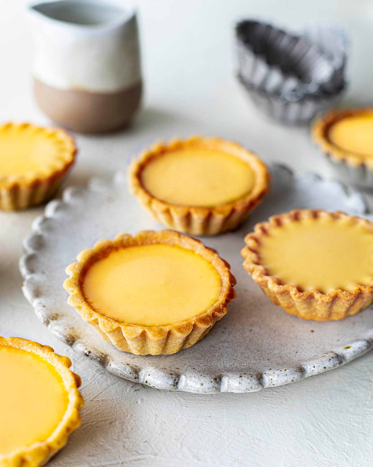 Multiple vegan egg tarts on a scalloped plate and around. Close up showing pretty crimped edges of one tart.