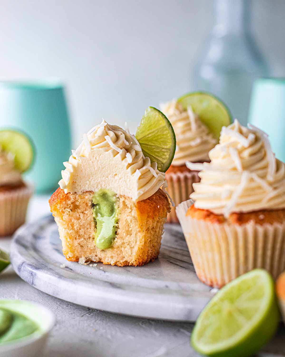 Vegan coconut lime cupcakes on marble board. One cupcake has a bite taken out of it showing the pastel green lime curd filling.