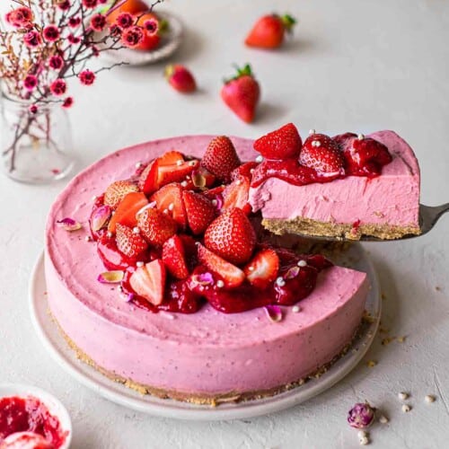 Vegan strawberry cheesecake on plate decorated with strawberry compote and fresh strawberries. Slice is lifted out on a cake server