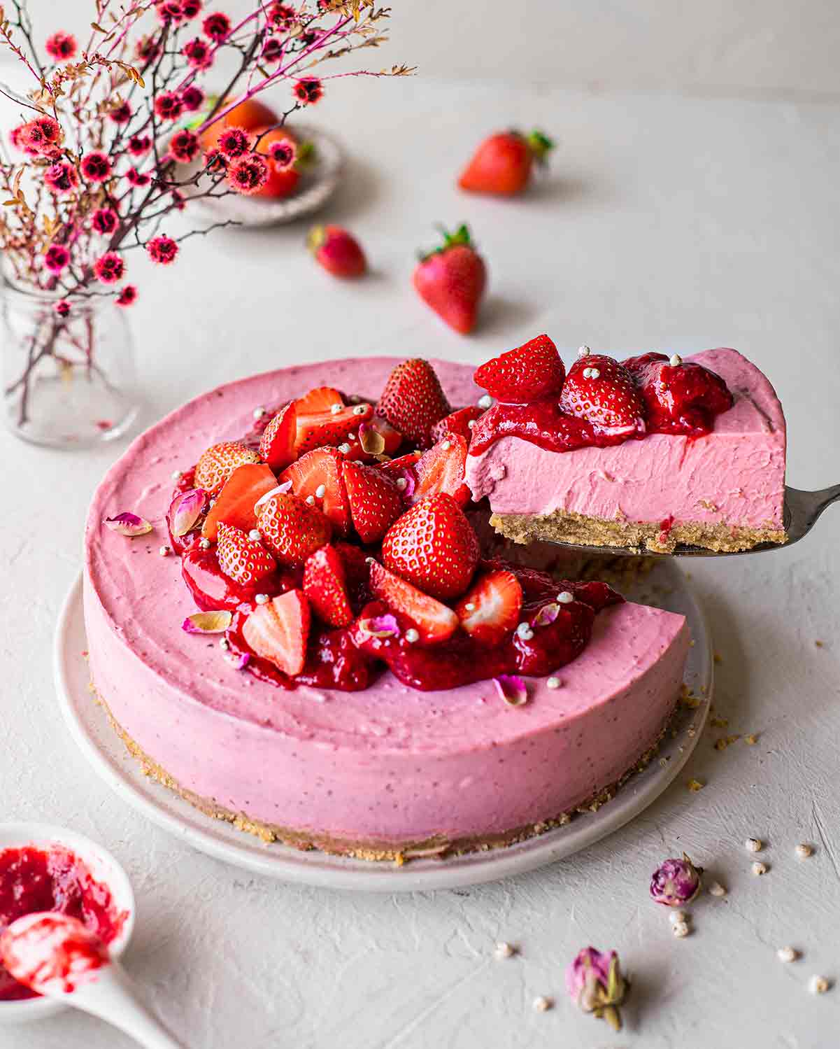 Vegan strawberry cheesecake on plate decorated with strawberry compote and fresh strawberries. Slice is lifted up on a cake server.