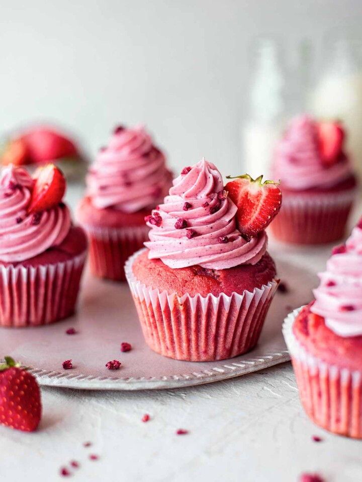 Vegan strawberry cupcakes with pink base and pink vegan buttercream. Cupcakes are decorated with freeze dried raspberries and fresh strawberries