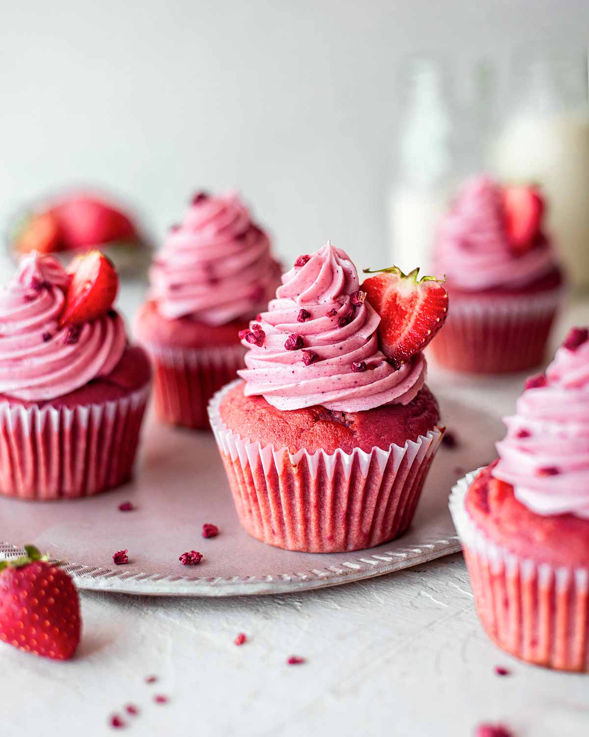 Vegan strawberry cupcakes on a serving plate with cupcakes in the foreground and background. Each cupcake is pink and topped with pink icing.