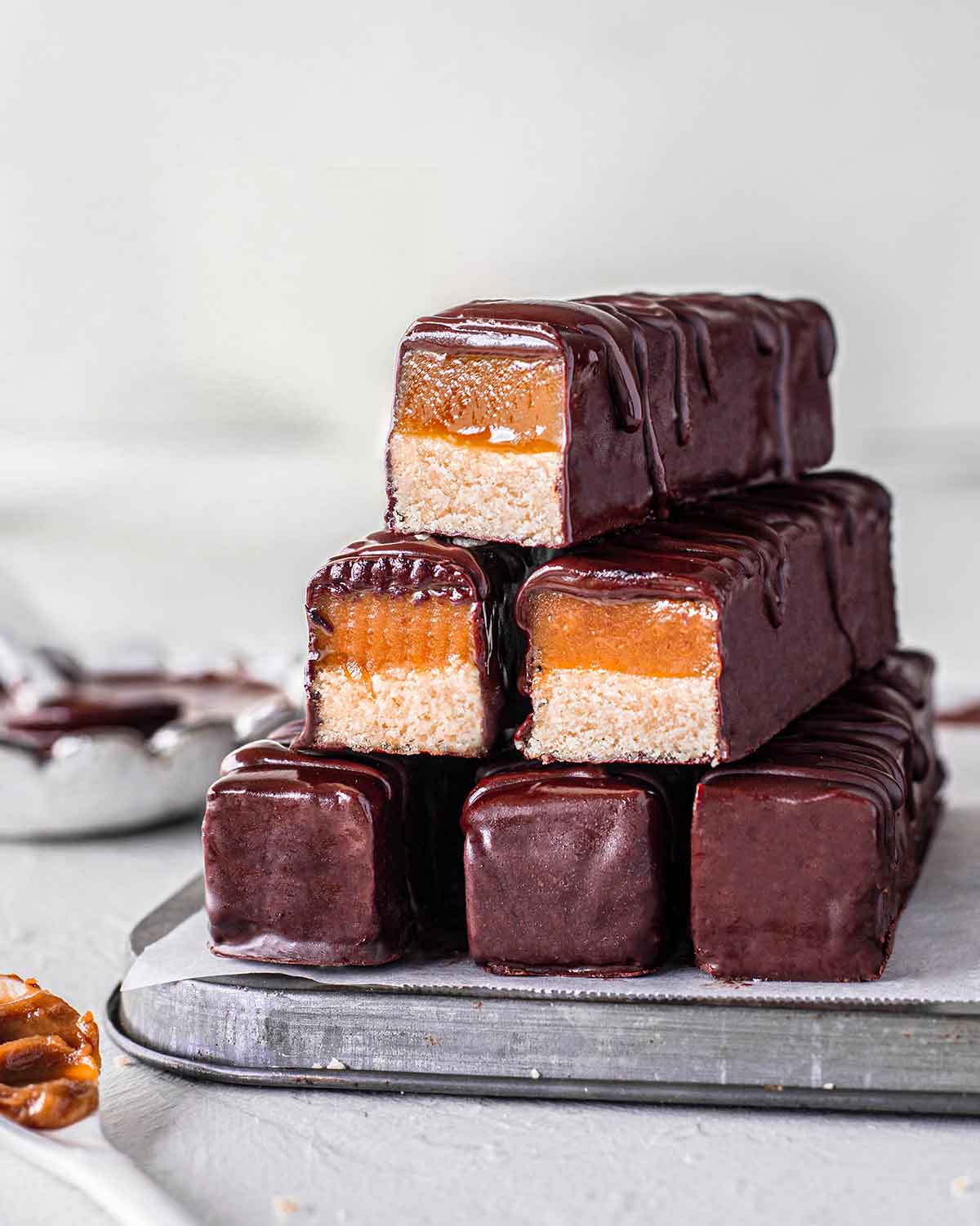 Stack of vegan twix bars on baking tray with chocolate in background. Three of the twix are cut showing their gooey caramel and cookie base.