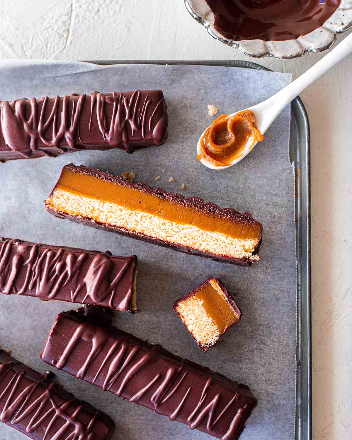 Flatlay of vegan twix bars on lined baking tray. One twix bar is sliced lengthways showing smooth caramel and golden cookie base.