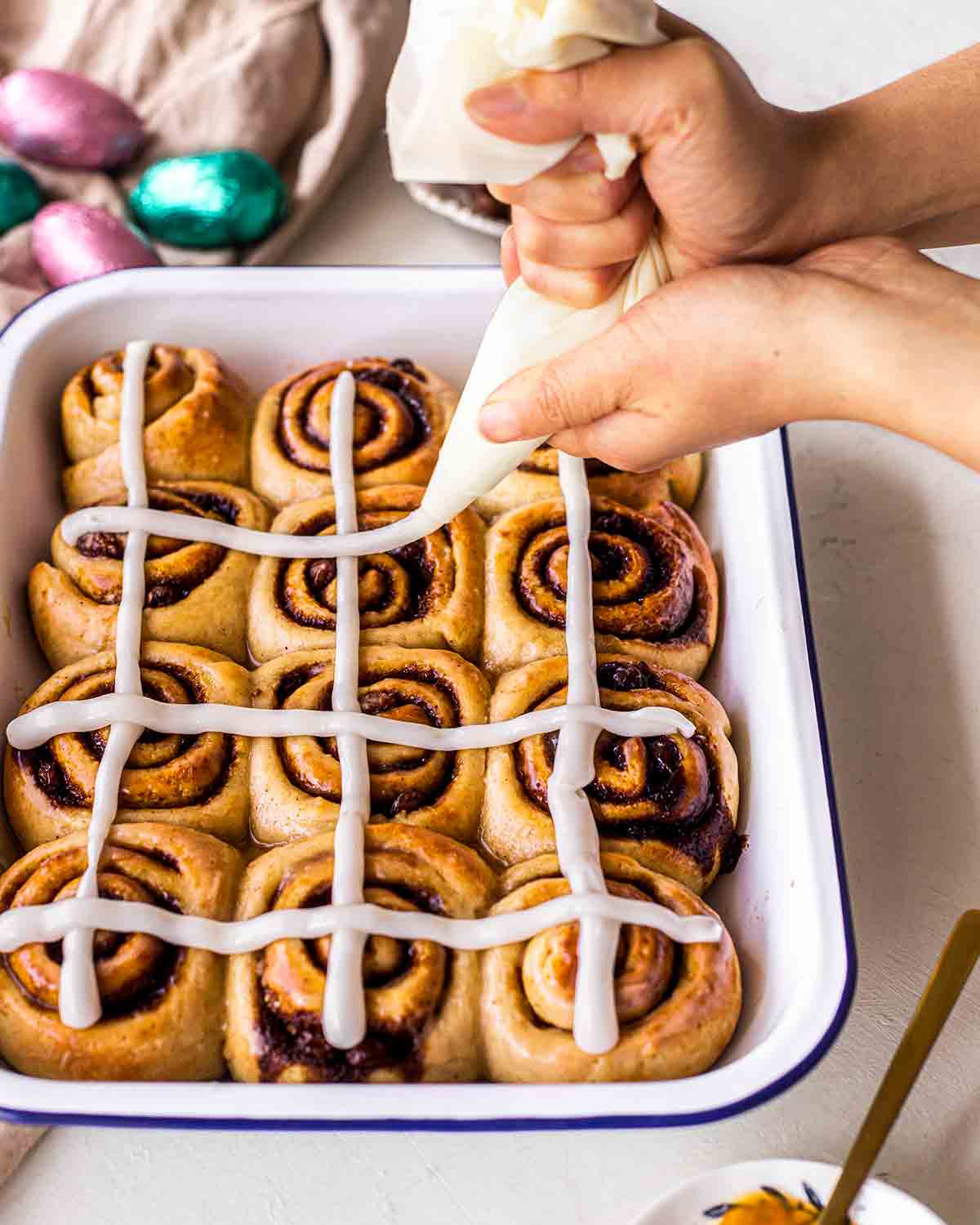 Vegan cinnamon rolls in a baking tray with two hands piping cream cheese frosting cross on the rolls.