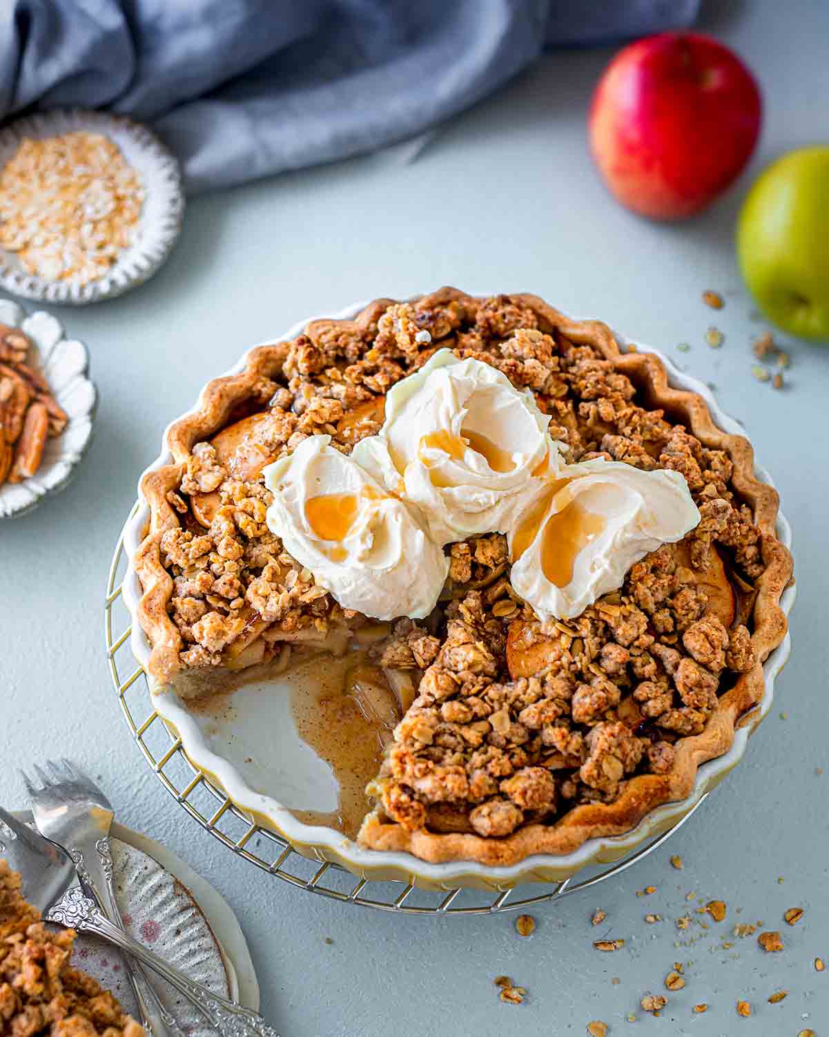 Vegan apple crumble pie in pie dish with a slice taken out. There are three dollops of thick cream on the pie with maple syrup poured on top. The filling is oozing out showing specks of cinnamon and nutmeg. The pie is surrounded by additional oat crumble, ingredients and a stack of dessert plates.