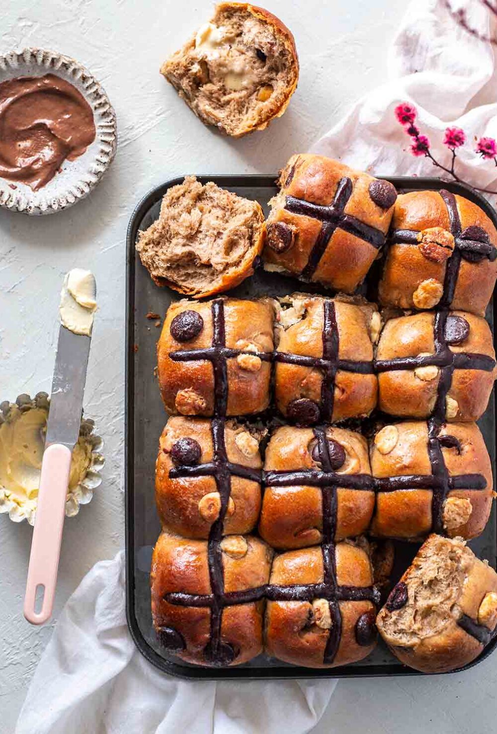 Double chocolate chip hot cross buns on baking tray. Two hot cross bun are cut open and has butter spread on one side. The other side is exposed showing soft bread and melting chocolate chips.