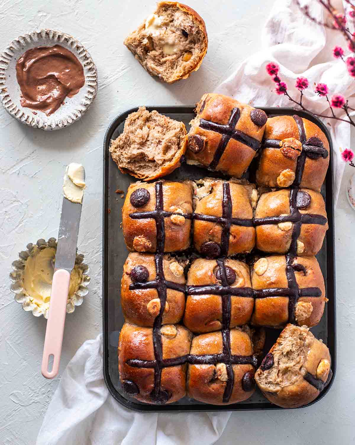 Double chocolate chip hot cross buns on baking tray. One hot cross bun is cut open and one side has vegan butter spread melting on it.
