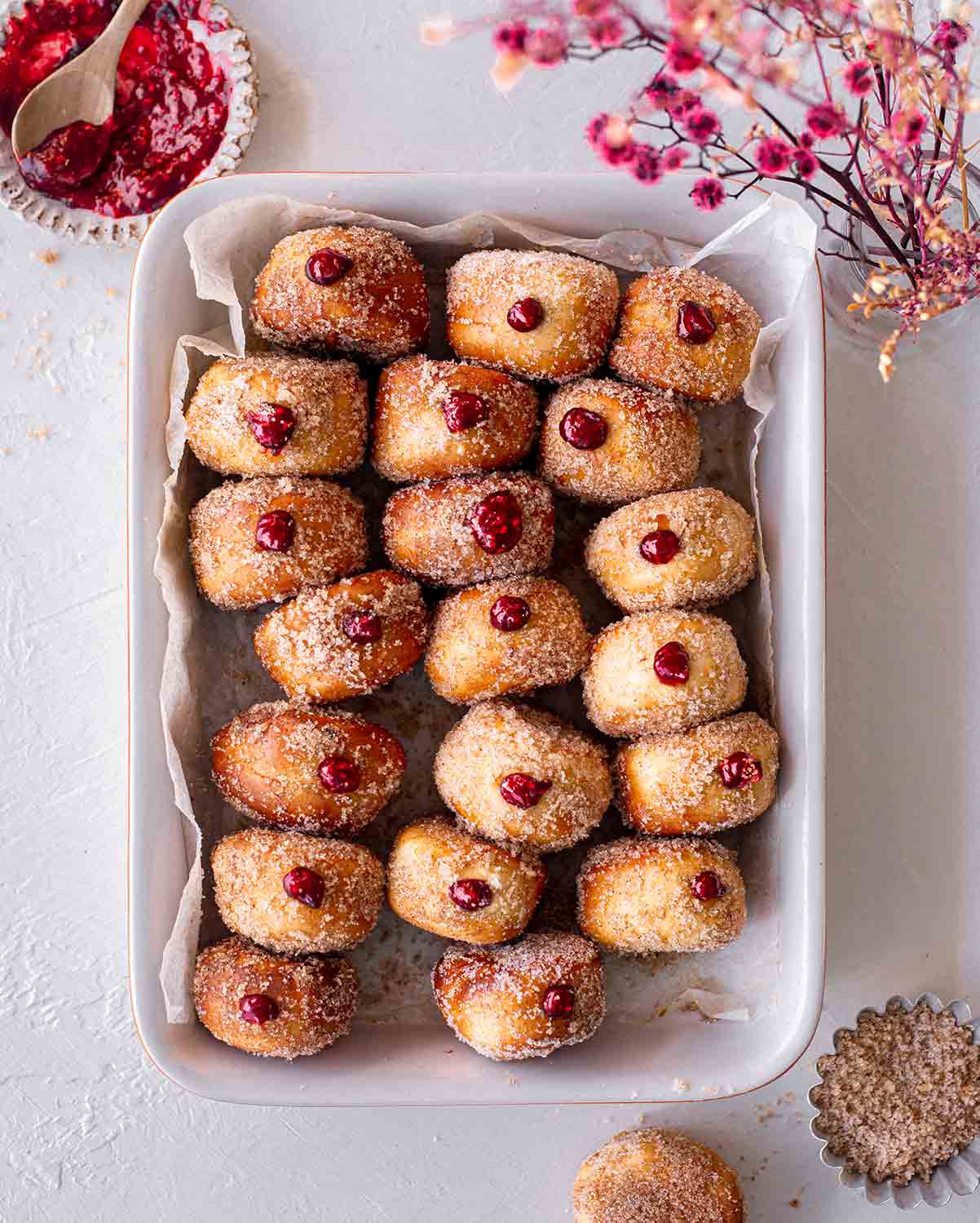 Lots of sugared mini donuts in a lined baking tray. Each donut has a little raspberry jam popping out.