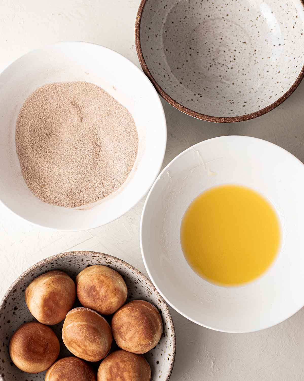 Flatlay of four bowls showing the undressed donuts, a bowl of melted vegan butter, a bowl of cinnamon sugar and an empty bowl for the final donuts.