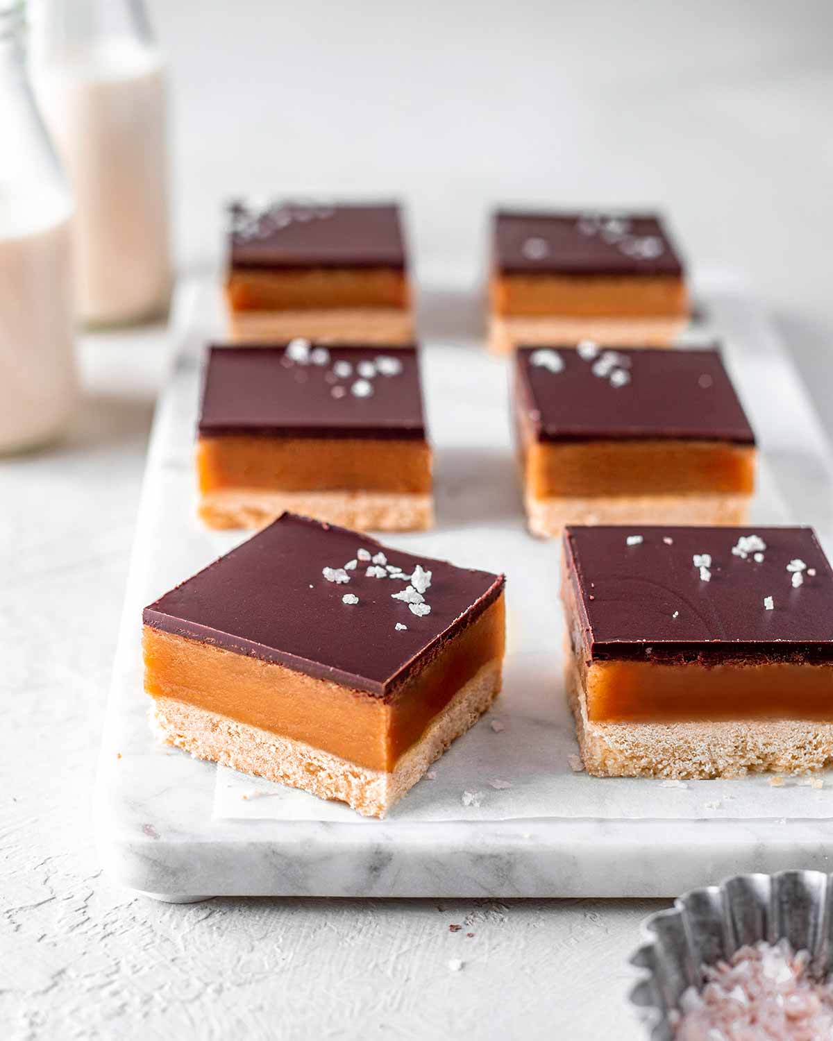 Vegan caramel slice or vegan millionaire bar on a white marble board. One slice is facing the light showing the even and smooth layers. Coarse sea salt is sprinkled on the chocolate layer of the slice.