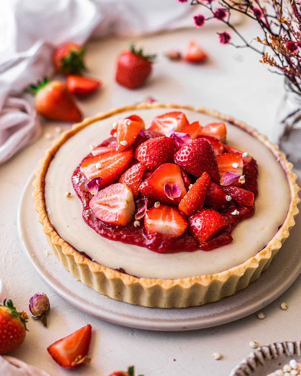 Vegan Strawberry Tart on plate. The tart has a buttery pale yellow shortcrust pastry, a thin coating of strawberry jam and is filled with a smooth pale cashew cream. The pie is then topped with strawberry compote and fresh berries.