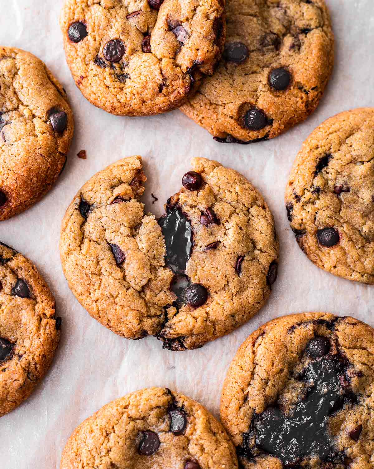 Flatlay of vegan chocolate chip cookies on a piece of baking paper. One cookie is broken in half revealing hot fudge centre.