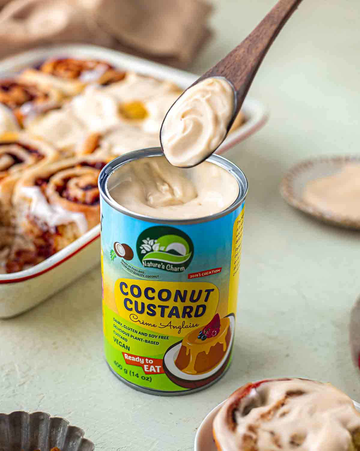 Nature's Charm coconut custard with a spoon coming out. The spoon is filled with creamy and thick custard. The vegan cinnamon rolls are in the background.