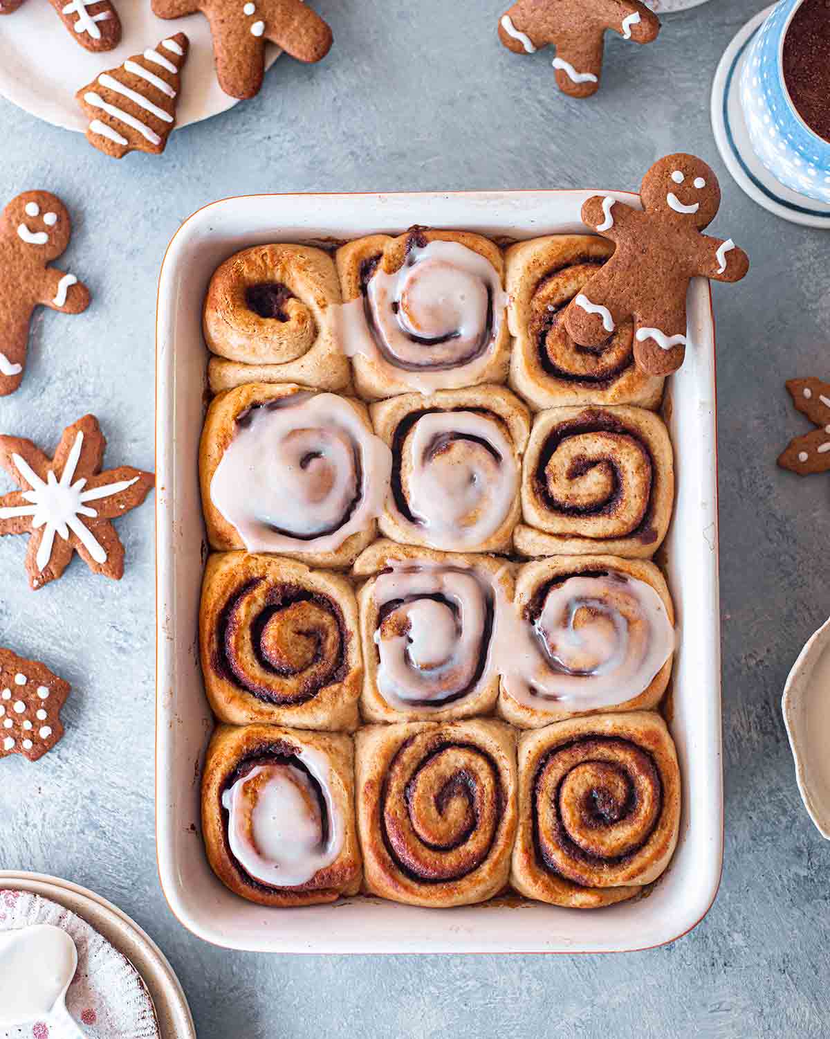 Vegan gingerbread cinnamon rolls in baking tray with gingerbread cut out cookies and hot chocolate surrounding it.