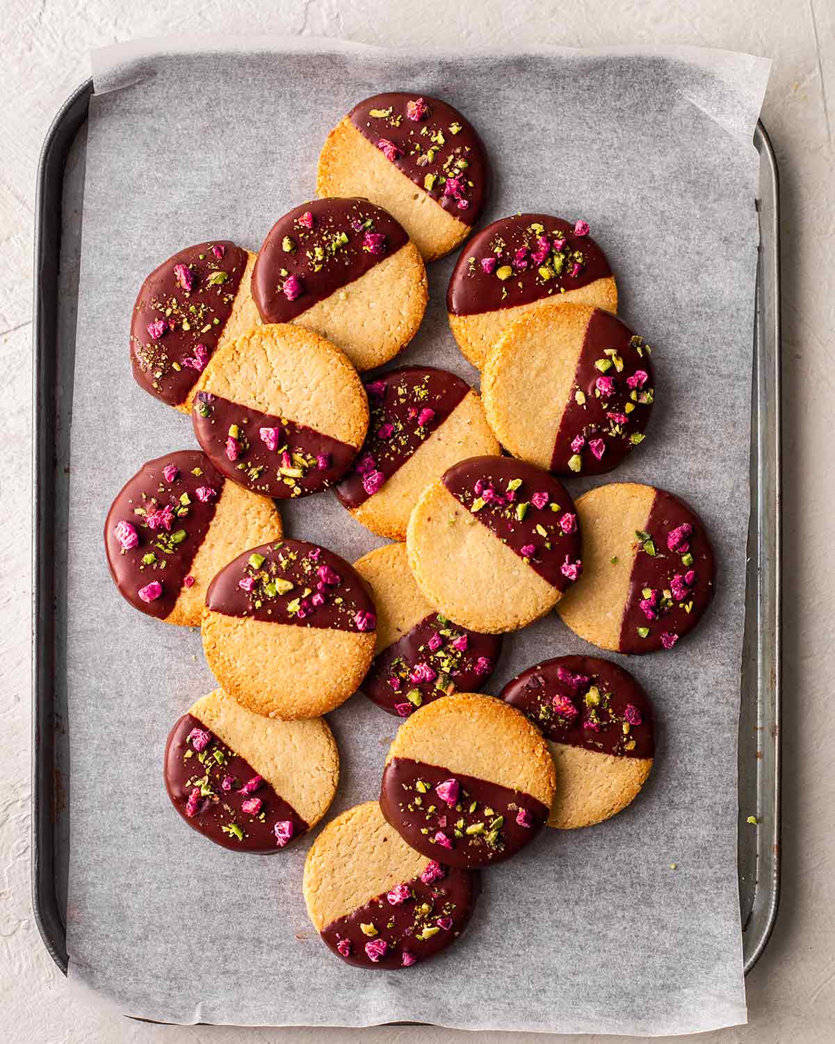 Almond flour cookies half dipped in chocolate with pistachios and freeze dried raspberries sprinkled on the chocolate. Cookies are on a lined baking tray.
