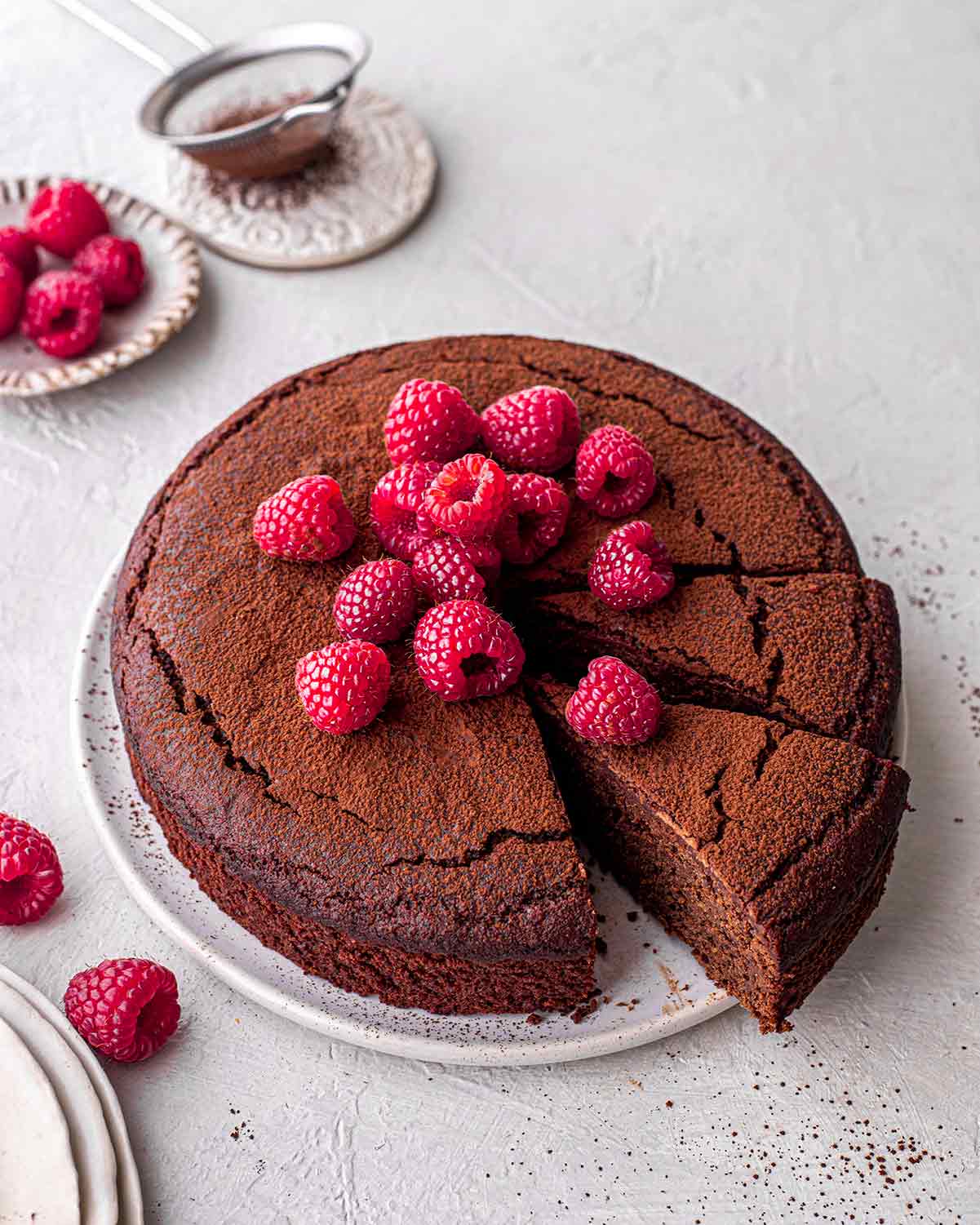 Flourless vegan gluten free chocolate cake on plate with a dusting of cocoa and fresh raspberries on top.