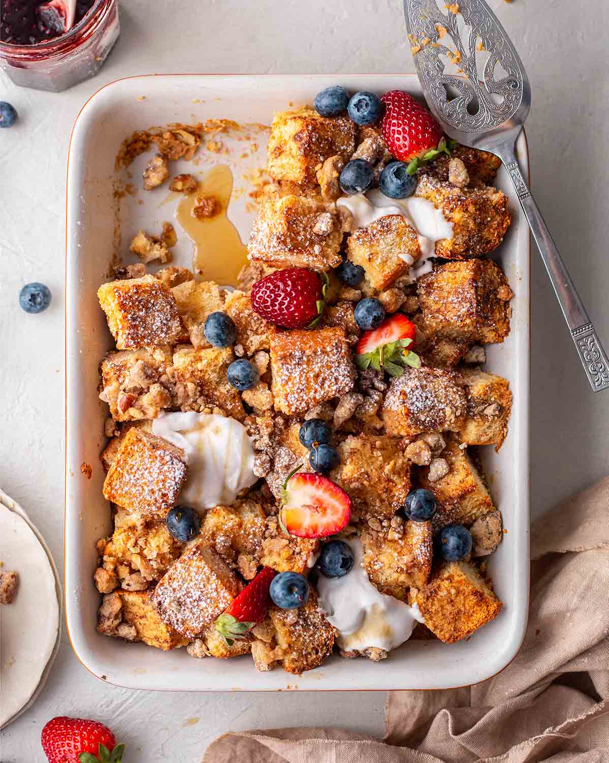 Vegan french toast cubes baked in a casserole dish. The dish is dusted with powdered sugar and is topped with a pecan crumble, berries and cream.