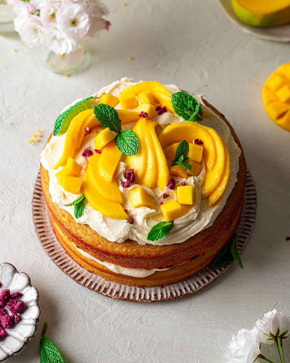 Decorated mango cake focusing on top decorated with cream cheese frosting, mango, fresh mint and freeze dried raspberries.