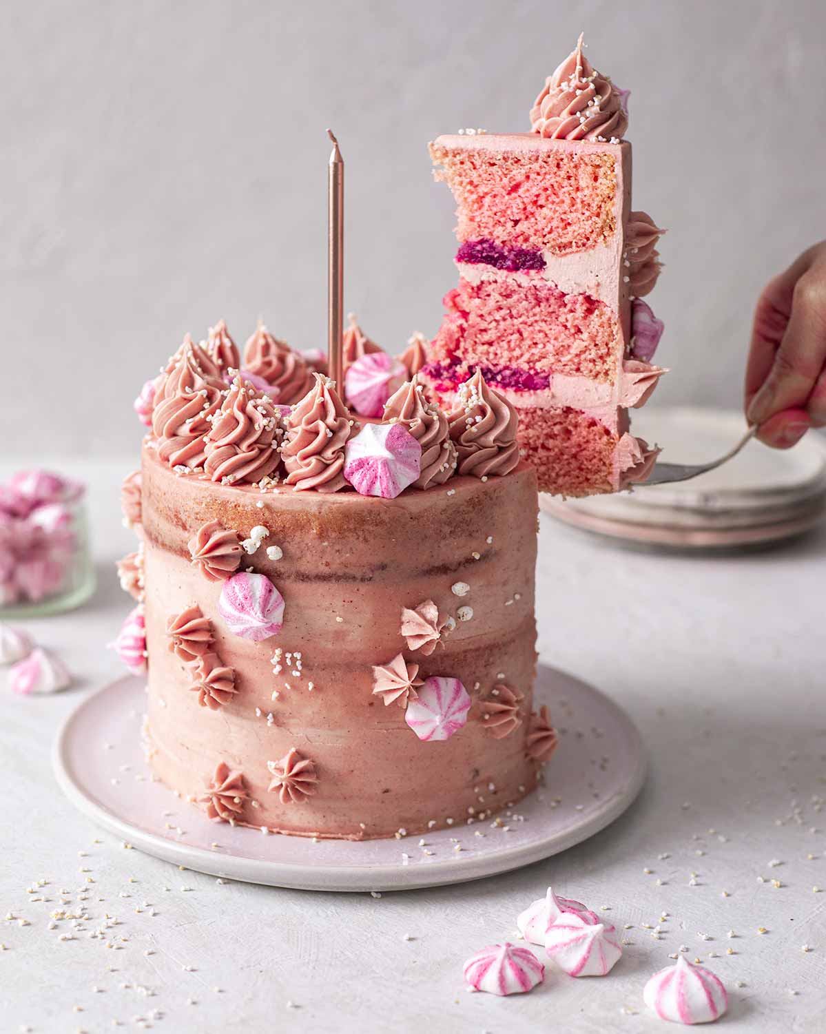 Pink vegan champagne cake decorated with pink buttercream, strawberry jam and meringue kisses. A slice is cut out of the cake and being lifted out.