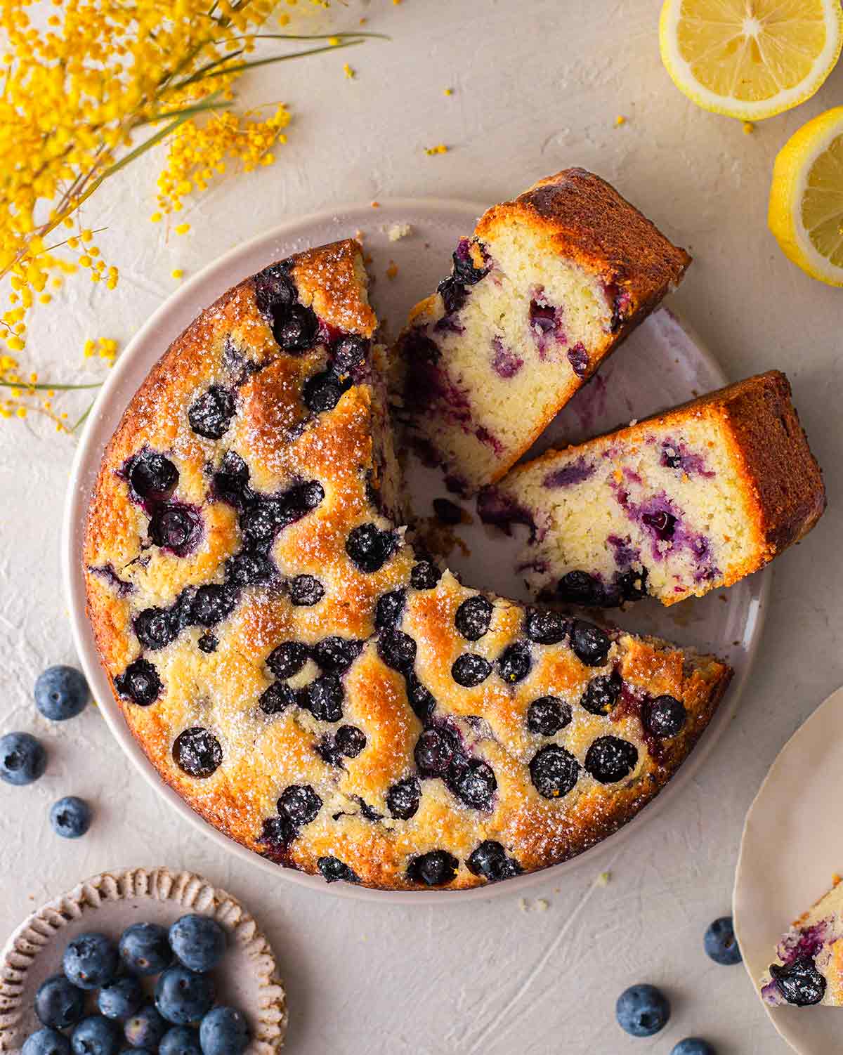 Vegan lemon blueberry cake on plate with two slices coming out showing fluffy golden texture.