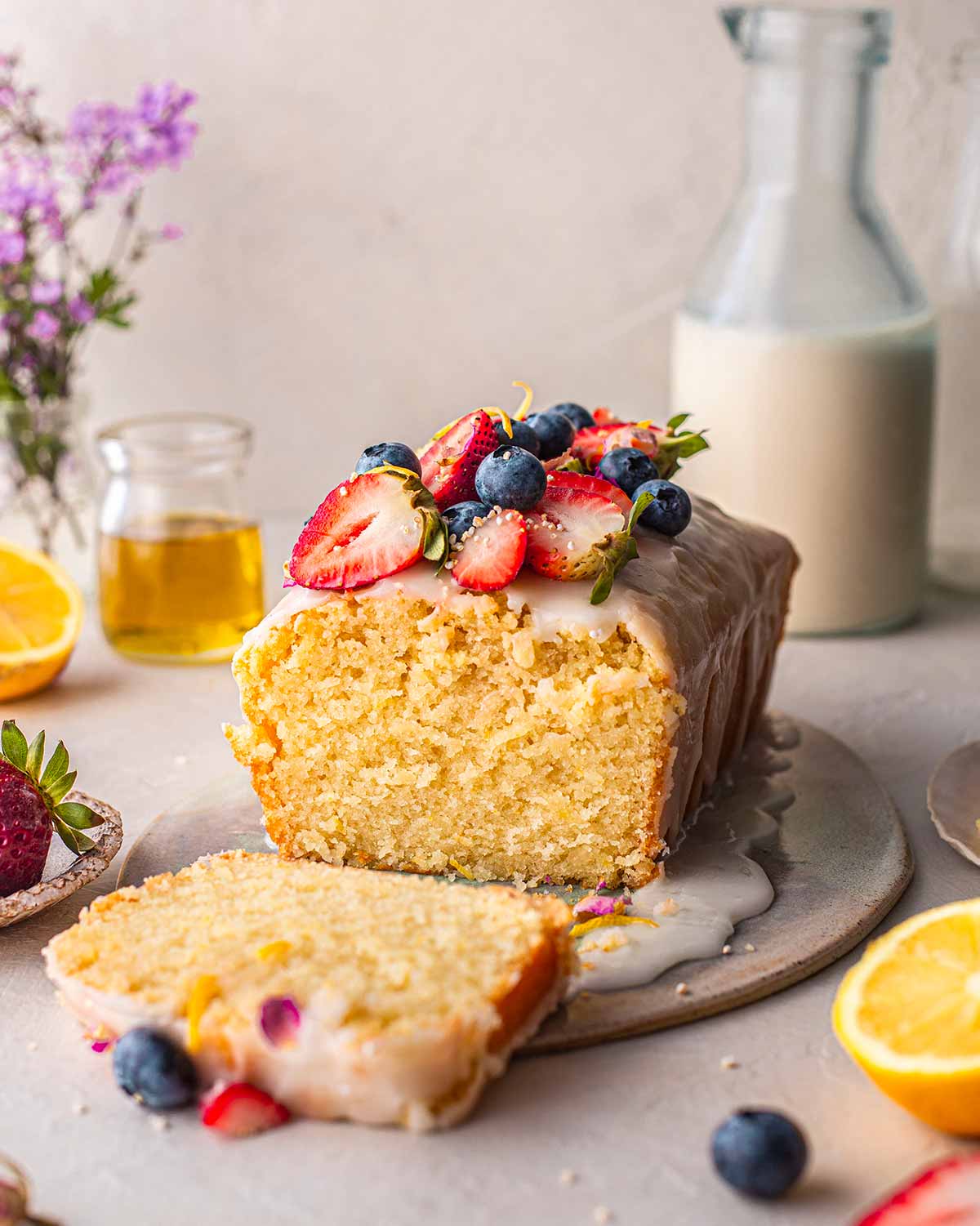 Lemon olive oil cake with a thin icing, blueberries and strawberries on top. Cake is on a plate amongst a few colourful items.