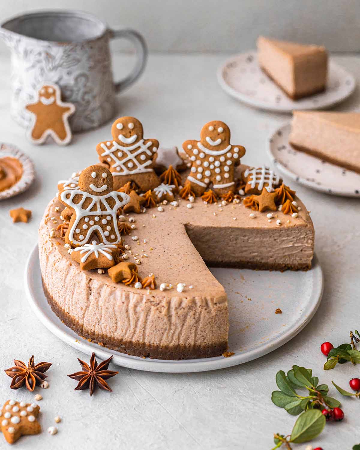 Vegan gingerbread cheesecake on plate with a few slices taken out showing creamy interior. Cheesecake is decorated with gingerbread cookies and piped Biscoff spread.
