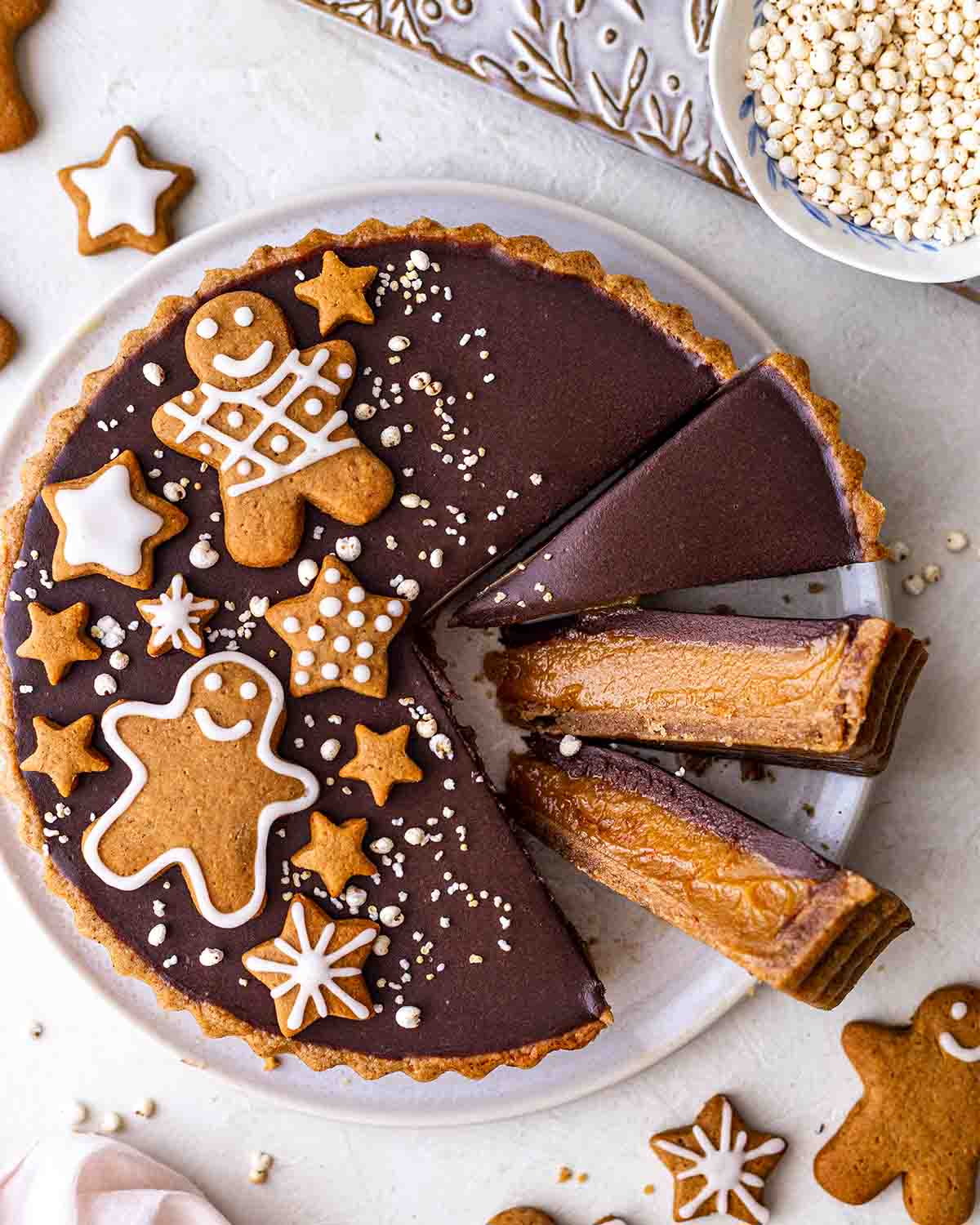 Vegan caramel tart with spiced crust and chocolate topping decorated with gingerbread cookies. The tart is on a plate with a few slices coming out showing gooey filling.