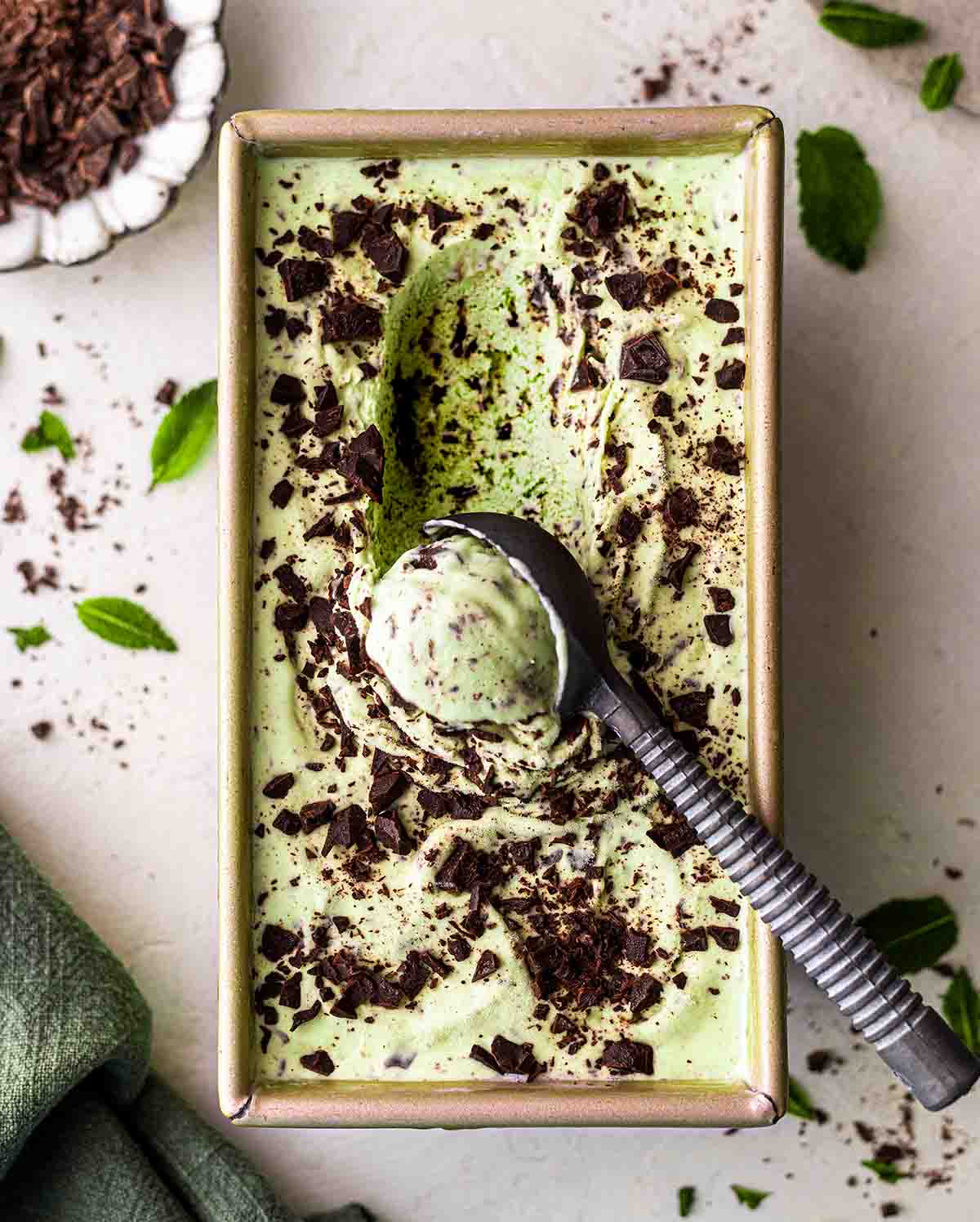 Creamy mint green ice cream in a loaf pan with lots of chopped chocolate throughout. An ice cream scoop is scooping away some ice cream showing soft texture.