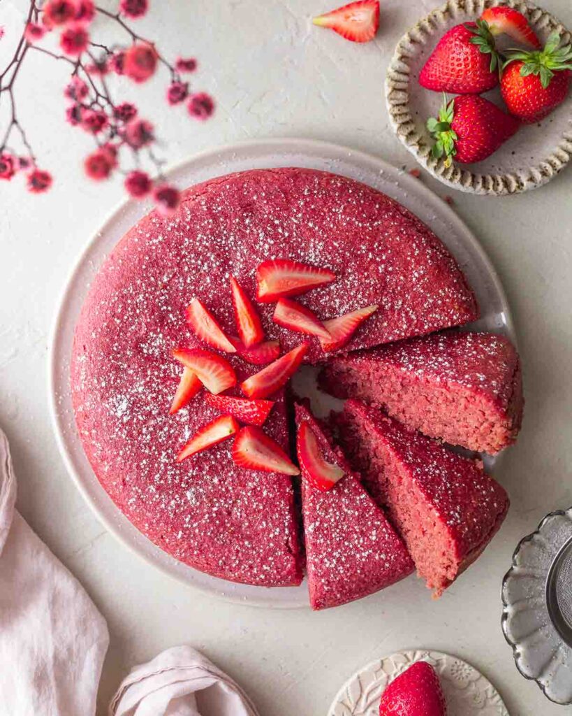 Overhead image of pink-colored vegan strawberry cake on a plate. A few slices are cut out showing fluffy texture.