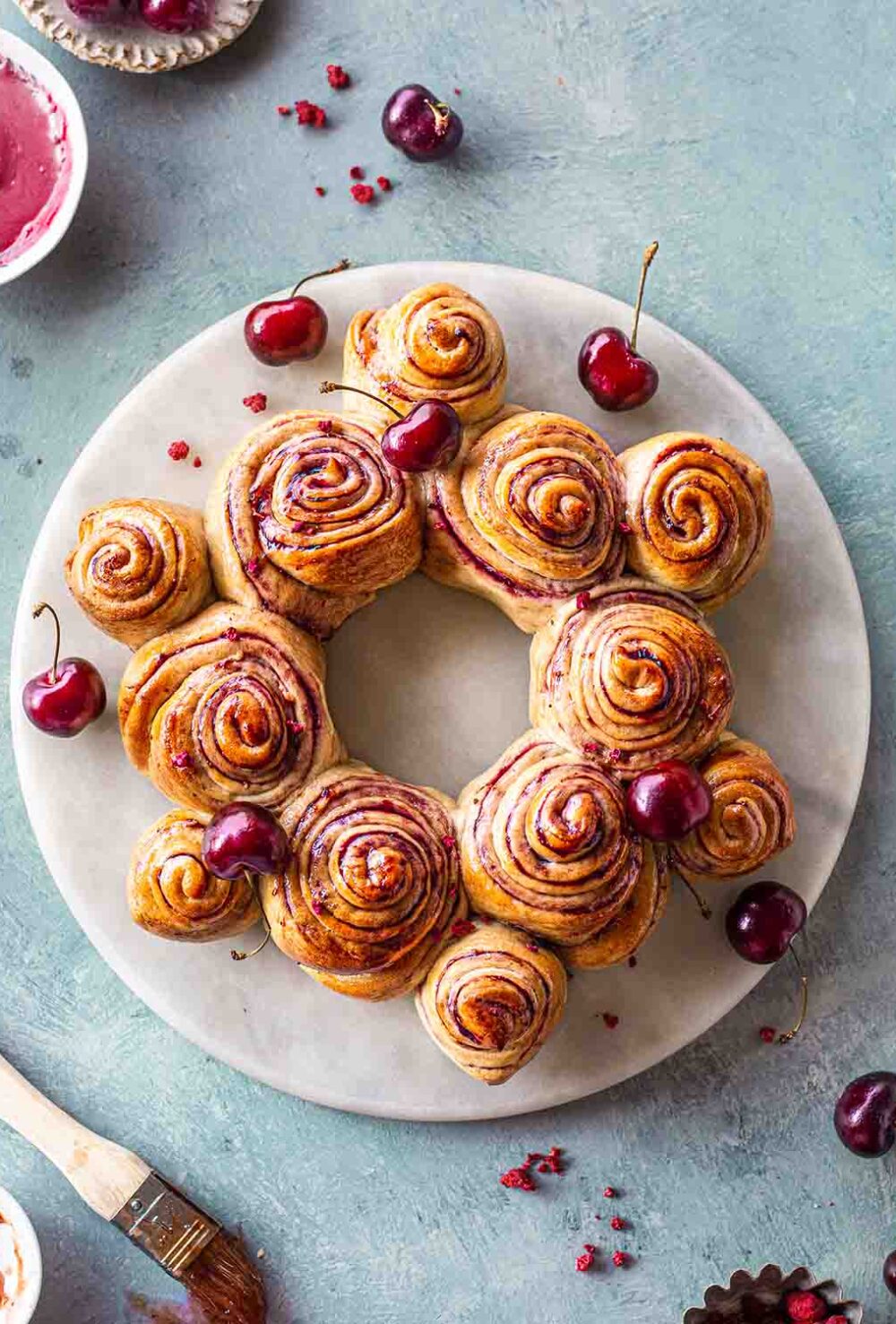 Cherry rolls shaped in a wreath on a marble serving platter. The wreath is surrounded by fresh cherries and condiments.