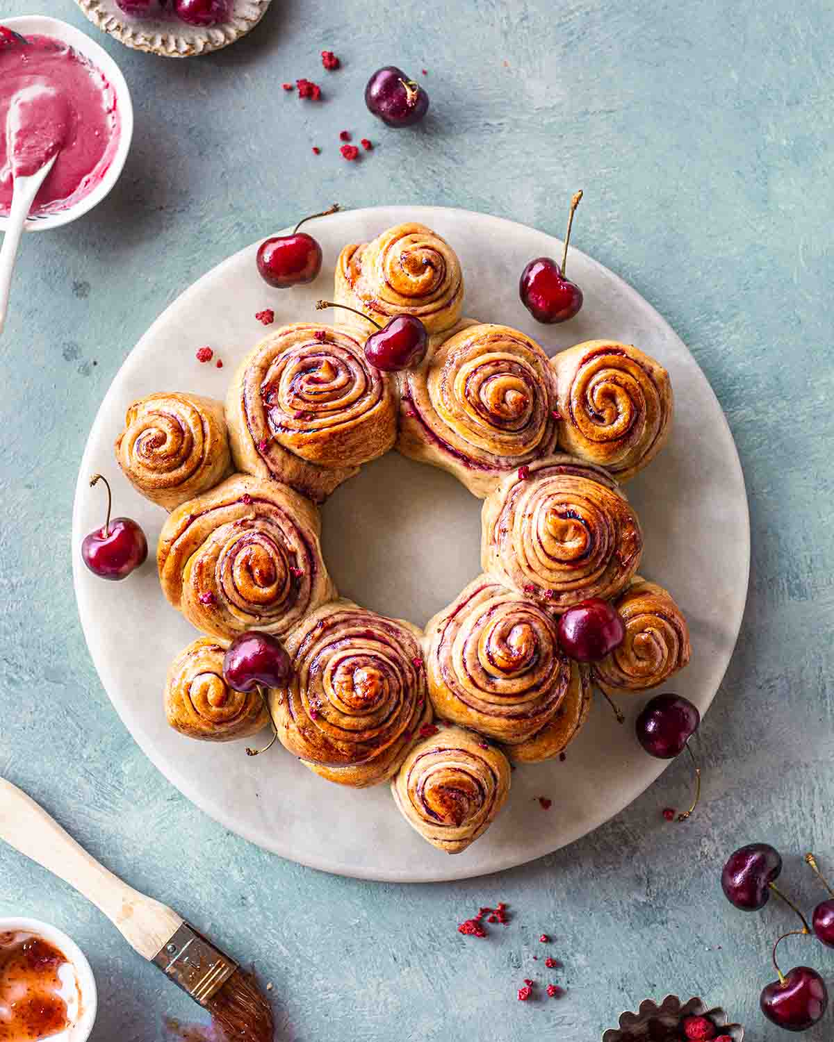Cherry rolls shaped in a wreath on a marble serving platter. The wreath is surrounded by fresh cherries and condiments.