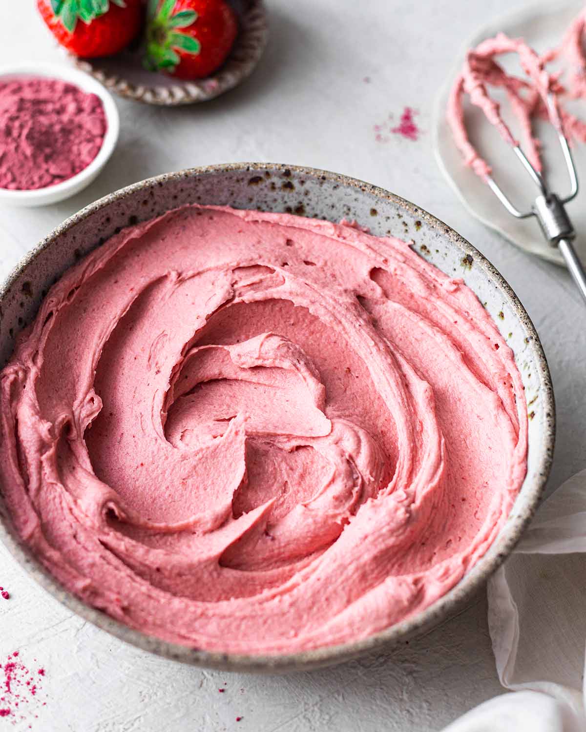 Bowl of creamy strawberry frosting with strawberry powder, fresh strawberries and hand beater whisk attachment in background.