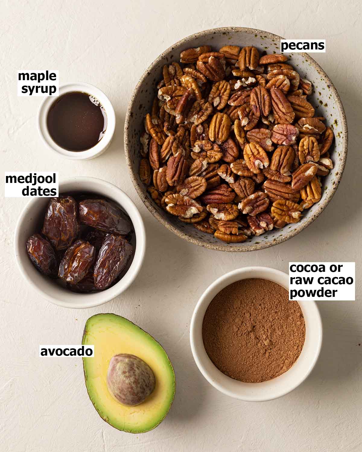 Flatlay of 5 ingredients including raw pecans, cocoa powder, medjool dates, avocado and maple syrup.